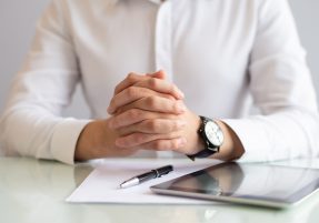 Close-up of male executive sitting at table with clasped hands. Caucasian hr manager working at table with papers and digital tablet. Employment concept